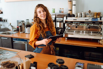 Smiling female barista takes an order from a panchette while standing at the bar counter in a...