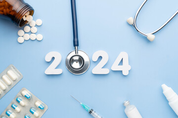 2024 Happy New Year banner for health care and medical concept. Stethoscope with doctor pills, syringe, vaccine vial, eyes drop, antibiotics and white number 2024 on table blue background. - obrazy, fototapety, plakaty