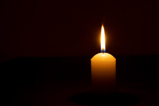 Single burning candle flame or light glowing on a big yellow candle on black or dark background on table in church for Christmas, funeral or memorial service with copy space.