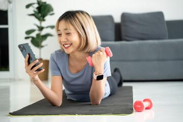 Smiling young woman with dumbbells and smartphone on yoga mat at home