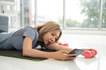 Lazy young woman with dumbbell and smartphone on yoga mat indoors