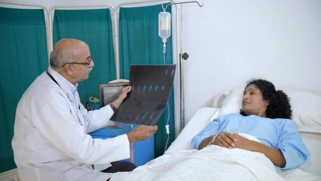 Indian old male doctor checking X-ray or MRI report for his female patient - radiology  CT-scan  X-ray image. A middle-aged Indian lady lying in the hospital bed - consulting a physician  medical p...