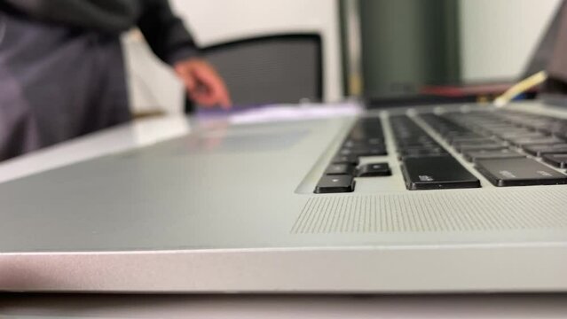 Close up view silver laptop keyboard black buttons with blurred unidentified male standing beside desk swipe and scroll phone screen on table. Digital and technology concept 