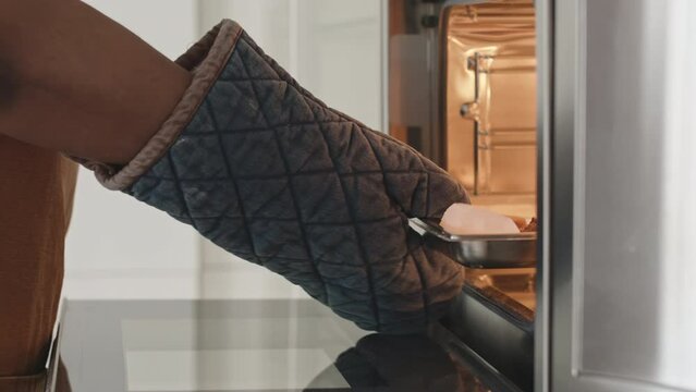 Close up impersonal shot of hands of African American housewife in mitt opening oven and taking off tray with baked cookies while preparing food in kitchen