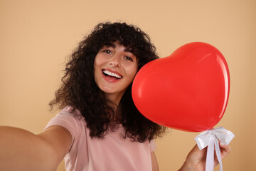 Happy young woman holding red heart shaped balloon and taking selfie on beige background
