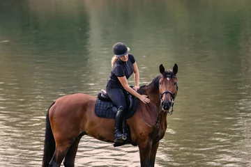 Foto op Canvas Female horseback rider in a black jockey outfit riding a chestnut horse along the river at sunset. Recreation, equitation, and nature concept. © 24K-Production