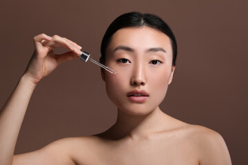 Beautiful young woman applying cosmetic serum onto her face on brown background