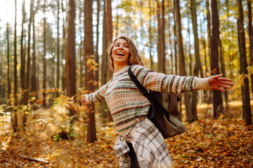 Tourist with a hiking backpack, hat walks along a path in the autumn forest. Beautiful woman enjoys a sunny day in nature, feels freedom and breathes fresh air, explores nature. - obrazy, fototapety, plakaty