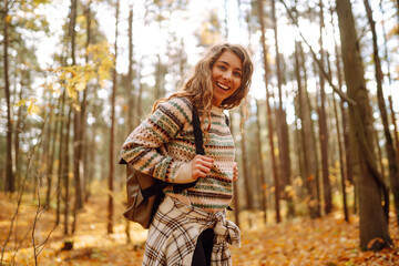 Tourist with a hiking backpack, hat walks along a path in the autumn forest. Beautiful woman enjoys...
