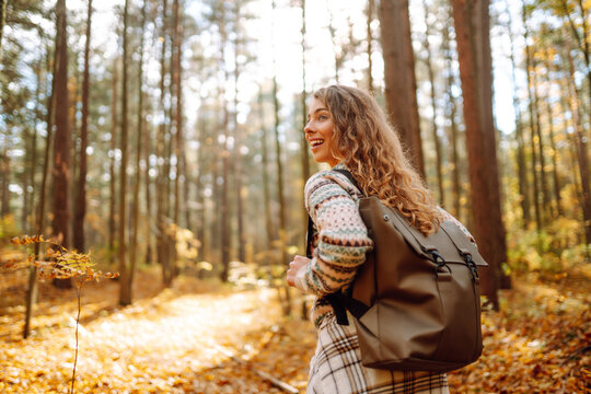 Tourist with a hiking backpack, hat walks along a path in the autumn forest. Beautiful woman enjoys a sunny day in nature, feels freedom and breathes fresh air, explores nature.