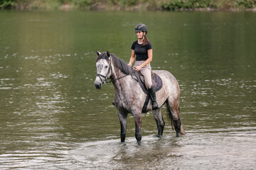 Gray horse with a female rider in the saddle walking through river water. Ecotourism and equestrian...