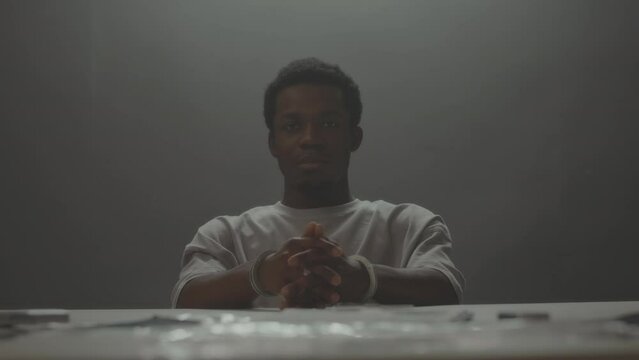 Zoom out portrait of arrested Black man looking frustratedly at camera sitting at desk in interrogation room with his hands locked in handcuffs