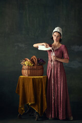 Elegant young woman in renaissance dress, maid pouring milk into glass against dark vintage...