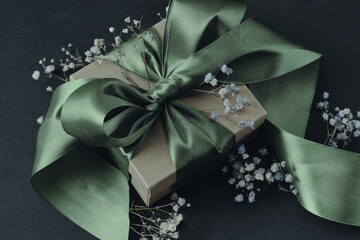 Paper gift box with olive green ribbon tied in a bow, small flowers, black background. Decoration for holidays, birthdays, weddings.