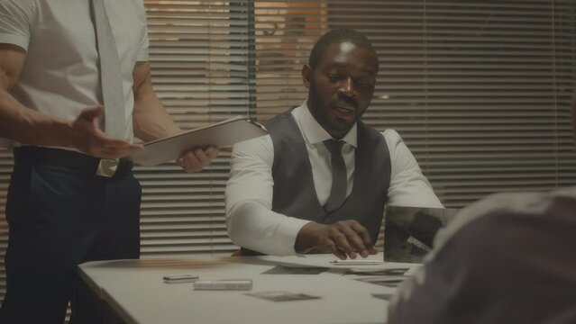 Medium shot of two diverse police officers interviewing African American criminal, showing pictures from crime scene to him in dark interrogation room