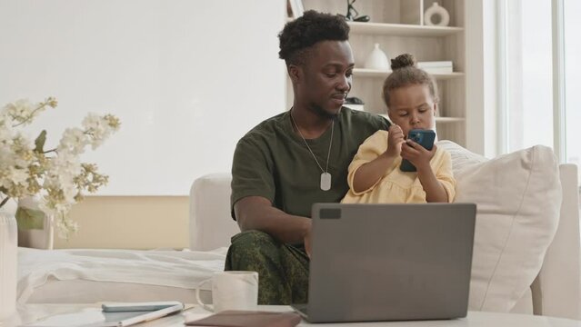 African American soldier in military uniform sitting with laptop on sofa in living room and playing with cute little daughter while spending day with family at home