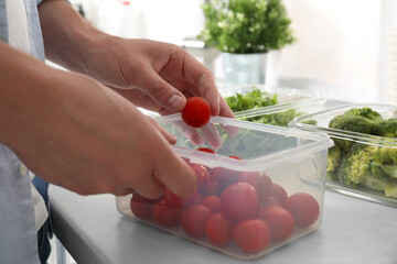 Man putting cherry tomato into plastic container with fresh vegetables in kitchen, closeup. Food...