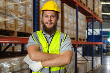 Bearded male industrial engineer in hard hat and safety suit arms crossed in car parts factory warehouse.