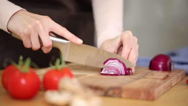Woman cutting red onion on wooden cutting board, slicing vegetables in bright sunny kitchen, vegetable scattered around