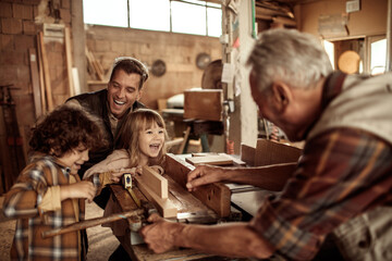 Grandfather carpenter teaching his son and grandchildren how to work with wood in a wood workshop - obrazy, fototapety, plakaty