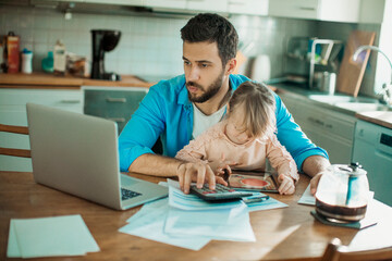 Young single father going over his bills and home finances while his daughter uses the tablet in...