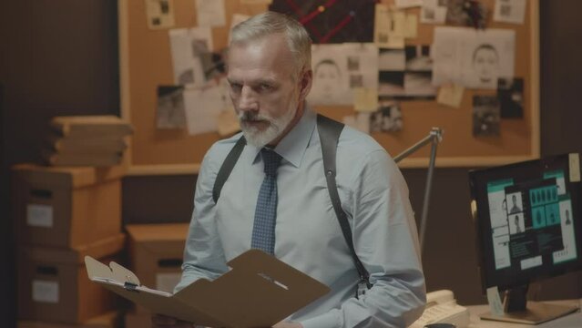 Medium portrait of mature Caucasian detective reading file folder with evidence of case investigation while working in dark office at night