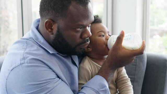 African Father Sitting On Sofa, Holding And Feeding Newborn Baby From Bottle Milk In Living Room. Authentic Lifestyle Candid Real Moment. Single Dad Family Home Life Concept.