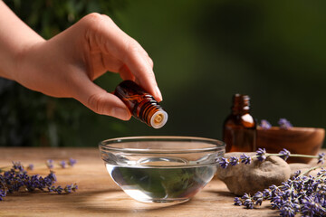 Woman dripping lavender essential oil from bottle into bowl at wooden table, closeup - obrazy, fototapety, plakaty