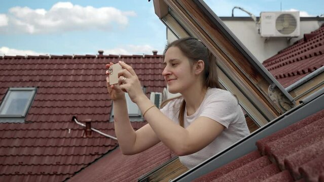 Young woman, with a bright smile, uses her smartphone to capture photos while looking out of an open attic window. Tourism, holidaying in Europe, and the spirit of travel and exploration.