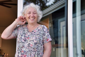 A happy elderly grey-haired woman smiles at the doorway of her house.