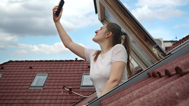 Young woman experiences signal issues with her cellphone as she peers out of an open attic window, attempting to regain a connection. Frustration of lost signal and connectivity problems.