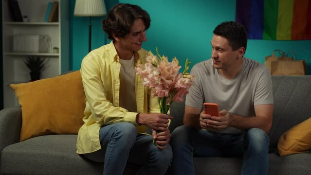 Video of a homosexual couple at home. One enters the frame with flower bouquet in hand, giving those to their partner, saying compliments, wishes, warm words.