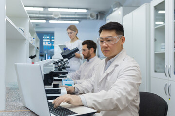 Fototapeta premium A young Asian man is concentrating on working on a laptop while sitting at a table in a research laboratory. A team of colleagues, a man and a woman working behind a microscope.
