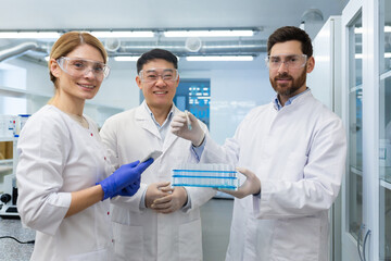 Portrait of an international group of chemists, doctors, scientists, men and women standing in a laboratory in uniforms and holding flasks with samples for research. They look and smile at the camera.