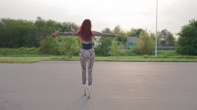 Slim Woman with Red Hair Has Fun Rollerblading on an Asphalt Parking Lot. Happy Girl Roller-Skates and Raises Her Arms to the Sides, Feeling Freedom and Wellbeing. Slow Motion.