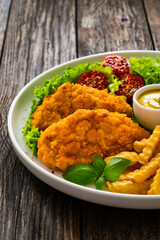 Seared breaded chicken nuggets with French fries and fresh vegetables on wooden table
