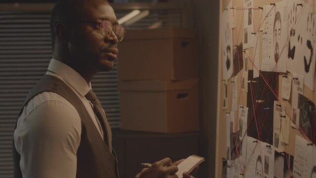 Tilt up shot of serious African American detective making notes while analyzing evidence board with suspects identikits, handprints and newspaper clippings connected with red thread