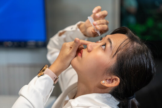 female Asian employee wearing glasses sits typing on a laptop and her eyesight hurts and she drops artificial tears into her eyes in a company meeting room.
