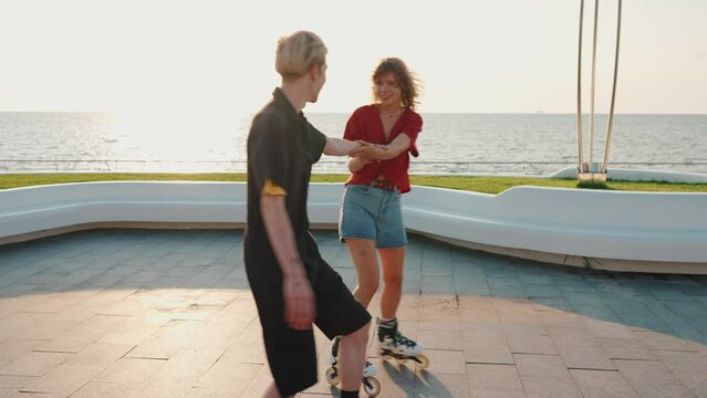 Cute young couple holding hands while rollerblading and hugging on sunset by the sea