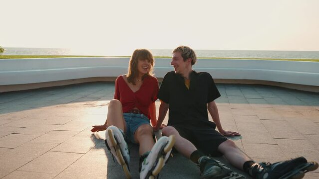 Laughing young couple talking while sitting on the floor after rollerblading on sunset by the sea