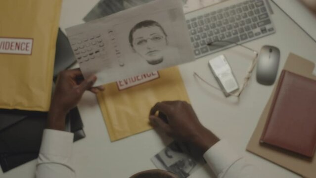 Top view of African American police detective looking at printed suspect identikit while investigating criminal case late in office