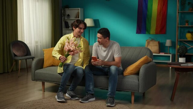 Full-size video of a homosexual couple at home. One enters the frame with flower bouquet in hand, giving those to their partner, saying compliments, wishes, warm words.