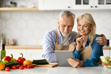 Smiling Senior Spouses Using Digital Tablet While Cooking Food In Kitchen Together
