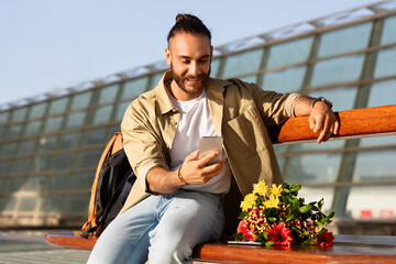 Positive millennial caucasian guy with bouquet waiting for wife, has video call on smartphone,...