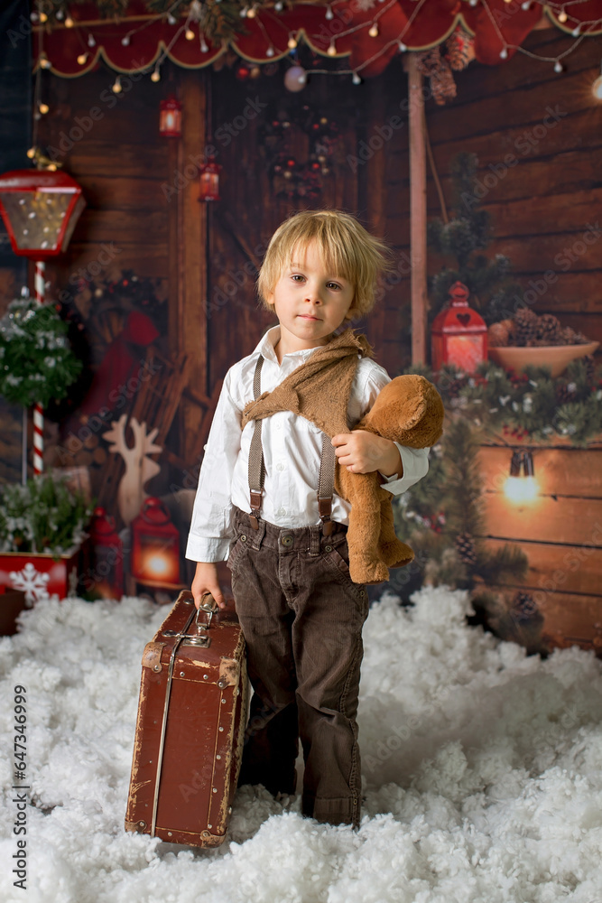 Wall mural Cute fashion toddler boy, playing in the snow with teddy bear in front of a wooden cabin log