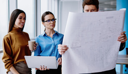 intelligent male and female colleagues standing in office looking at blueprint and accountings cooperating on project