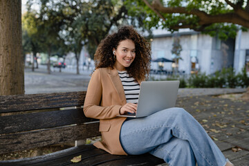 pretty curly woman sitting in city street in stylish jacket, working remote job on laptop