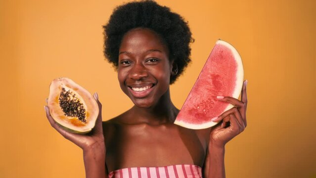 African American young woman with afro hair holding papaya and watermelon in hands isolated on orange background. Skin care and spa treatments.