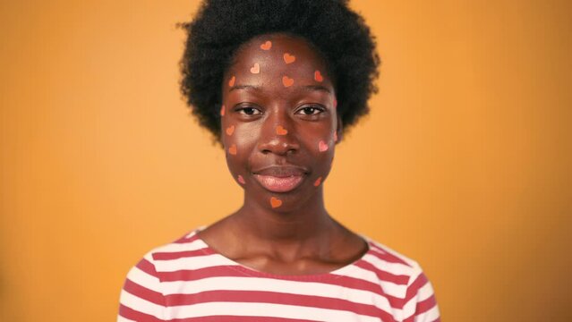 African American young woman with afro hair standing with small hearts pasted on her face isolated on orange background. Valentine's Day.