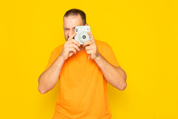 Bearded Hispanic man in his 40s wearing an orange t-shirt taking a photograph with an instant camera, isolated on yellow studio background.